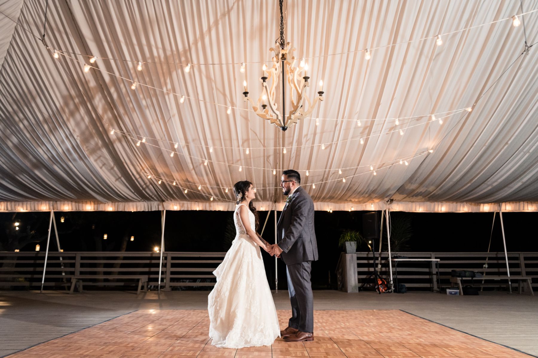 Bride and groom, hold hands underneath a bistro lit white tent with a chandelier above their head at Stonehouse Villa.