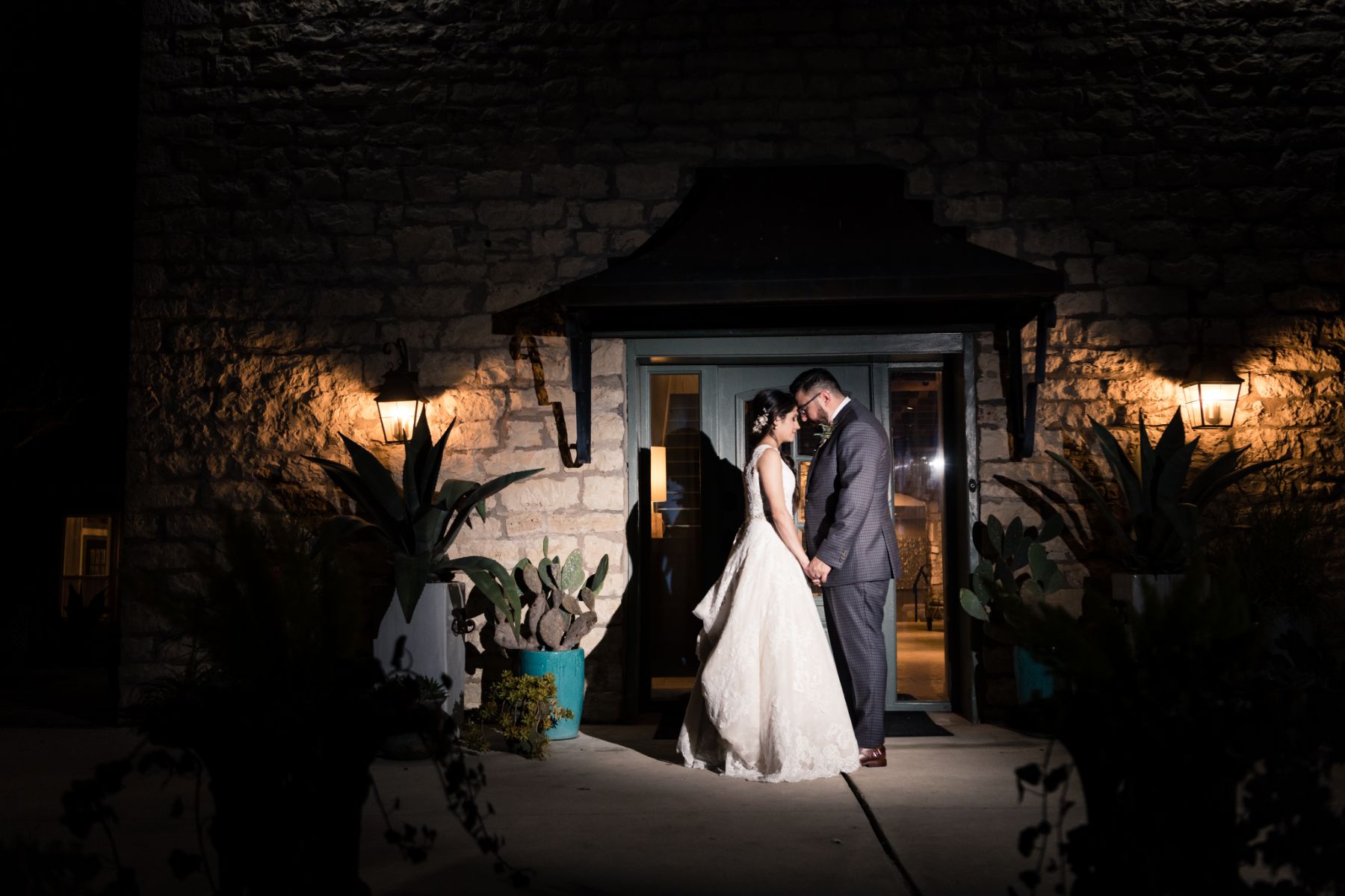 Bride and groom are holding hands with their foreheads touching, two lanterns are lit on each side with a limestone building behind them.