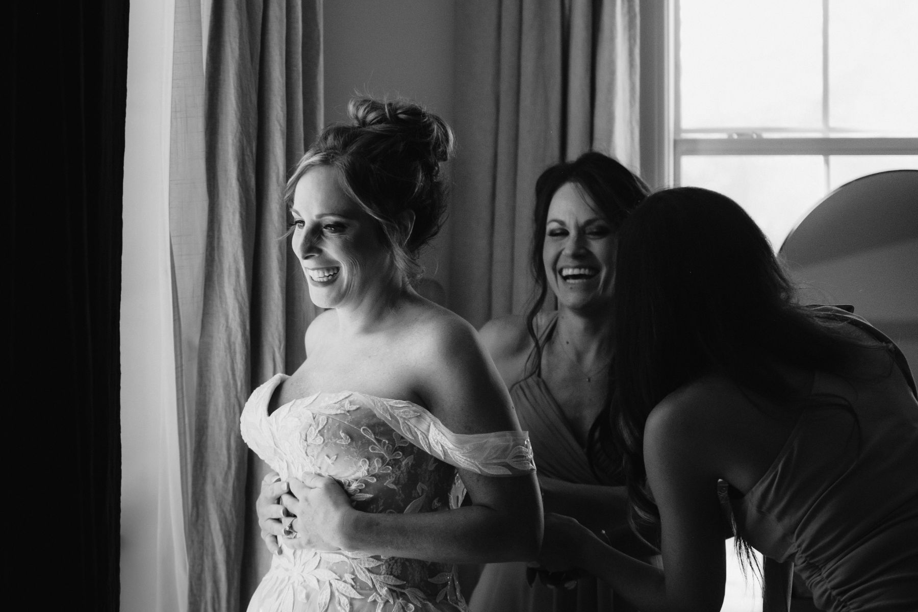 A bride smiles, while to bridesmaids help zip up her dress.
