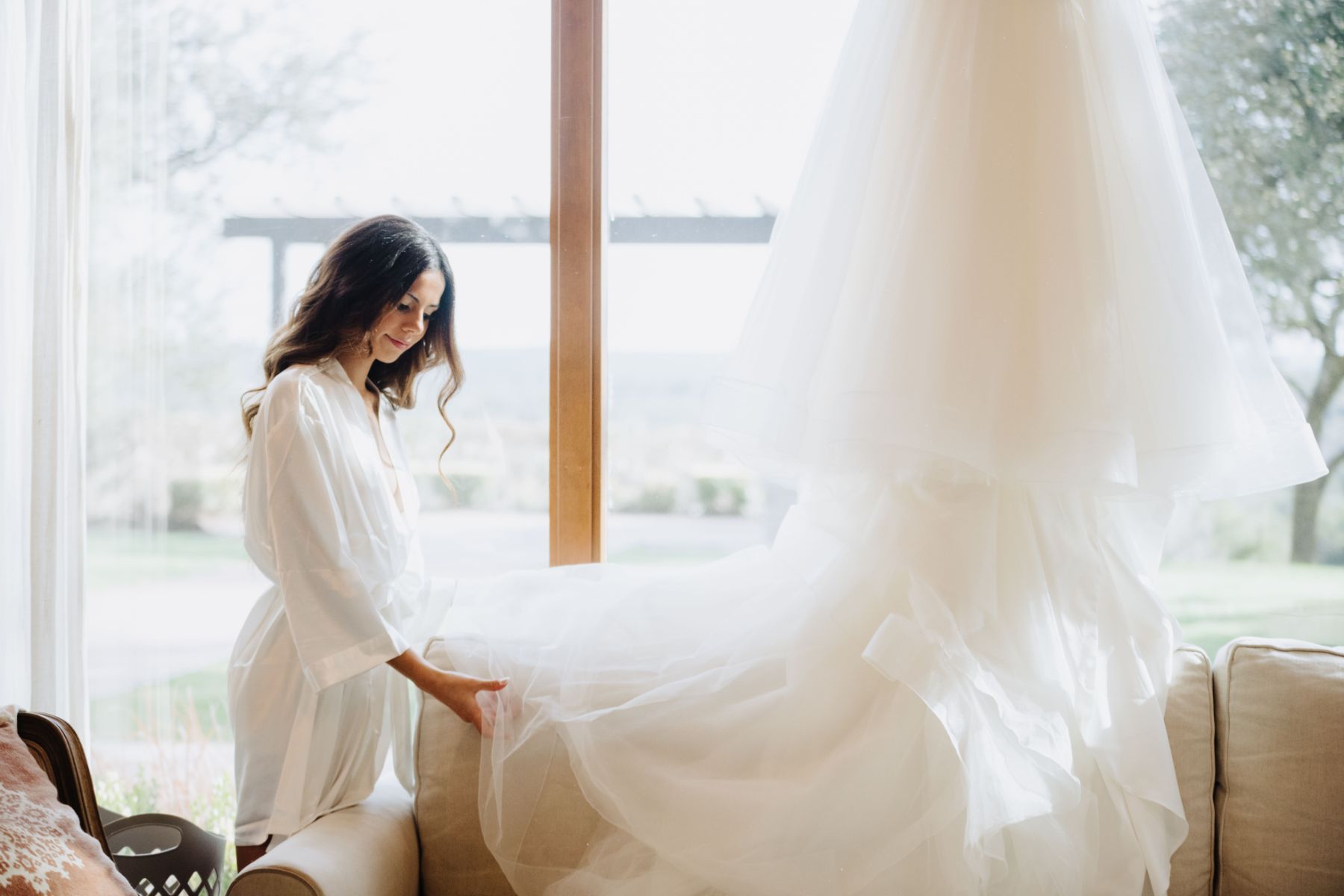 A bride is standing near a wedding dress hanging on a hook. The bride is touching the hem of the tool dress. Large windows are in the background that look out to the Texas Hill country.