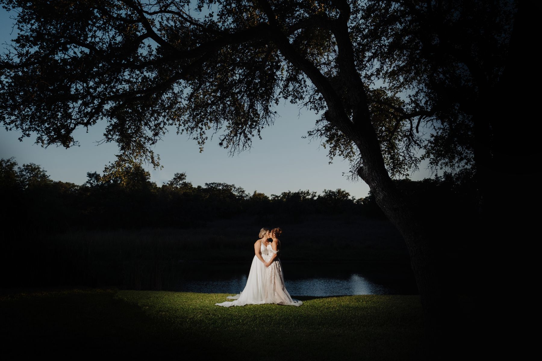 Two brides hold hands and kiss under large oak trees with the sun setting over the horizon and a pond in the background.