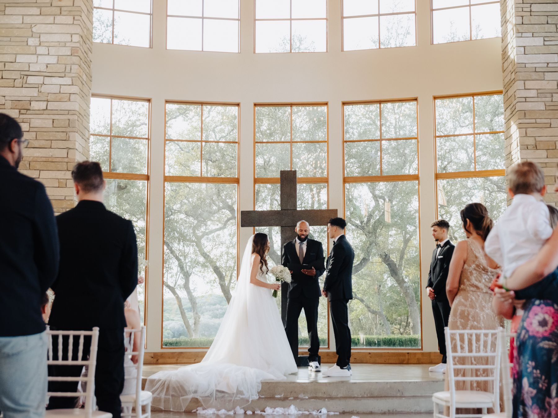 Bride and groom stand at wedding altar with a cross in the background. Texas oak trees can be seen through the large windows behind the couple at Canyonwood Ridge Wedding venue.