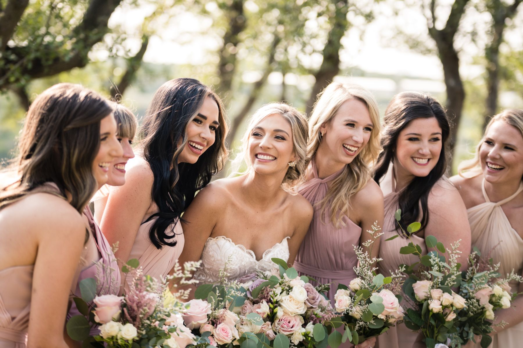 A bride is smiling and looking at her bridesmaids, whom are also smiling and laughing together. The sun is illuminating their hair for a soft serene image.