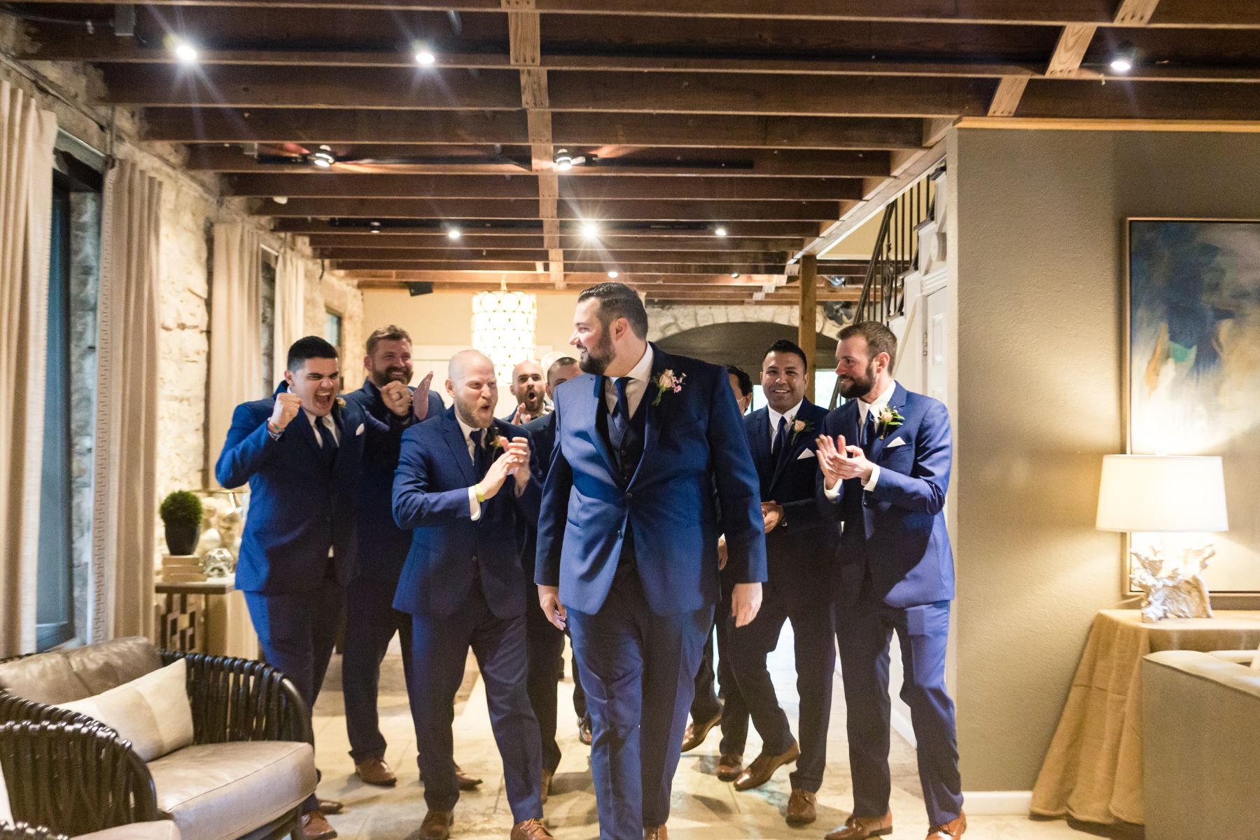 Groom walks with groomsmen as they cheer him on in a rustic, elegant room at Stonehouse Willa with exposed wood beams and limestone walls.