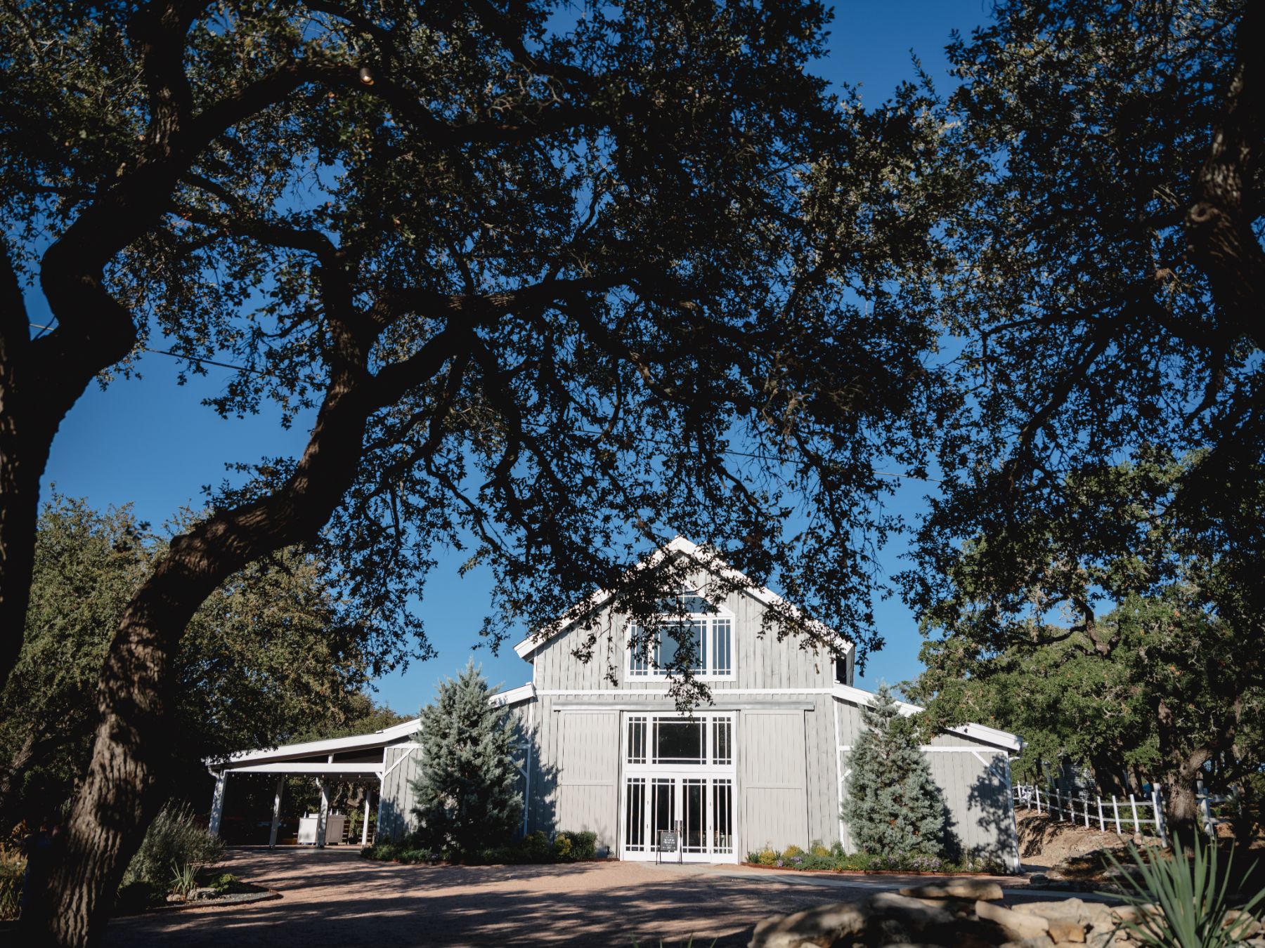 Large oak trees, and a blue sky frame, the gray barn of the Addison Grove wedding venue in Austin Texas.