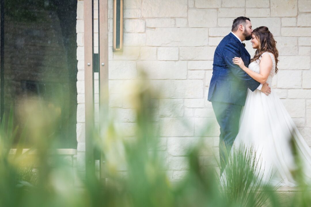 Groom, kisses bride on forehead with stone building in the background.