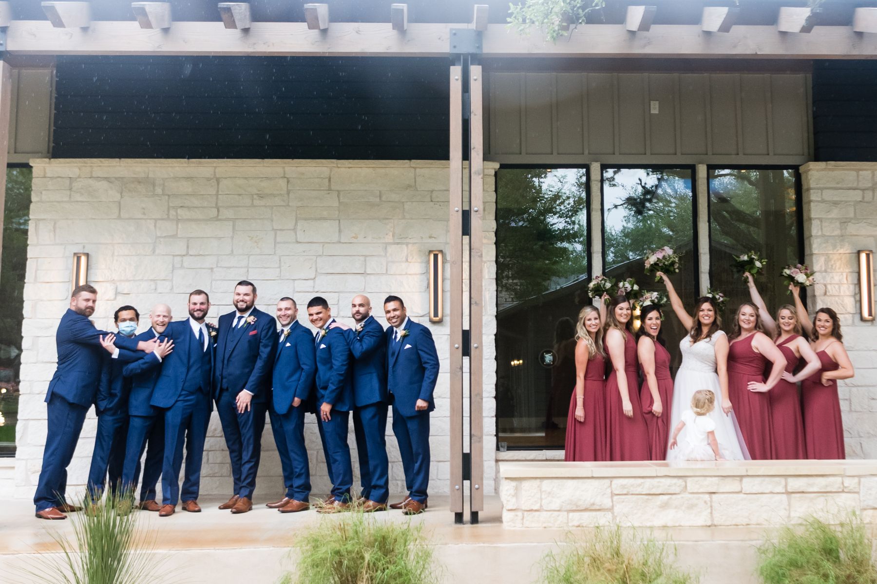 Groom and groomsmen stand on the left side with the bride and bridesmaids, standing on the right side. A limestone building and patio are behind them.