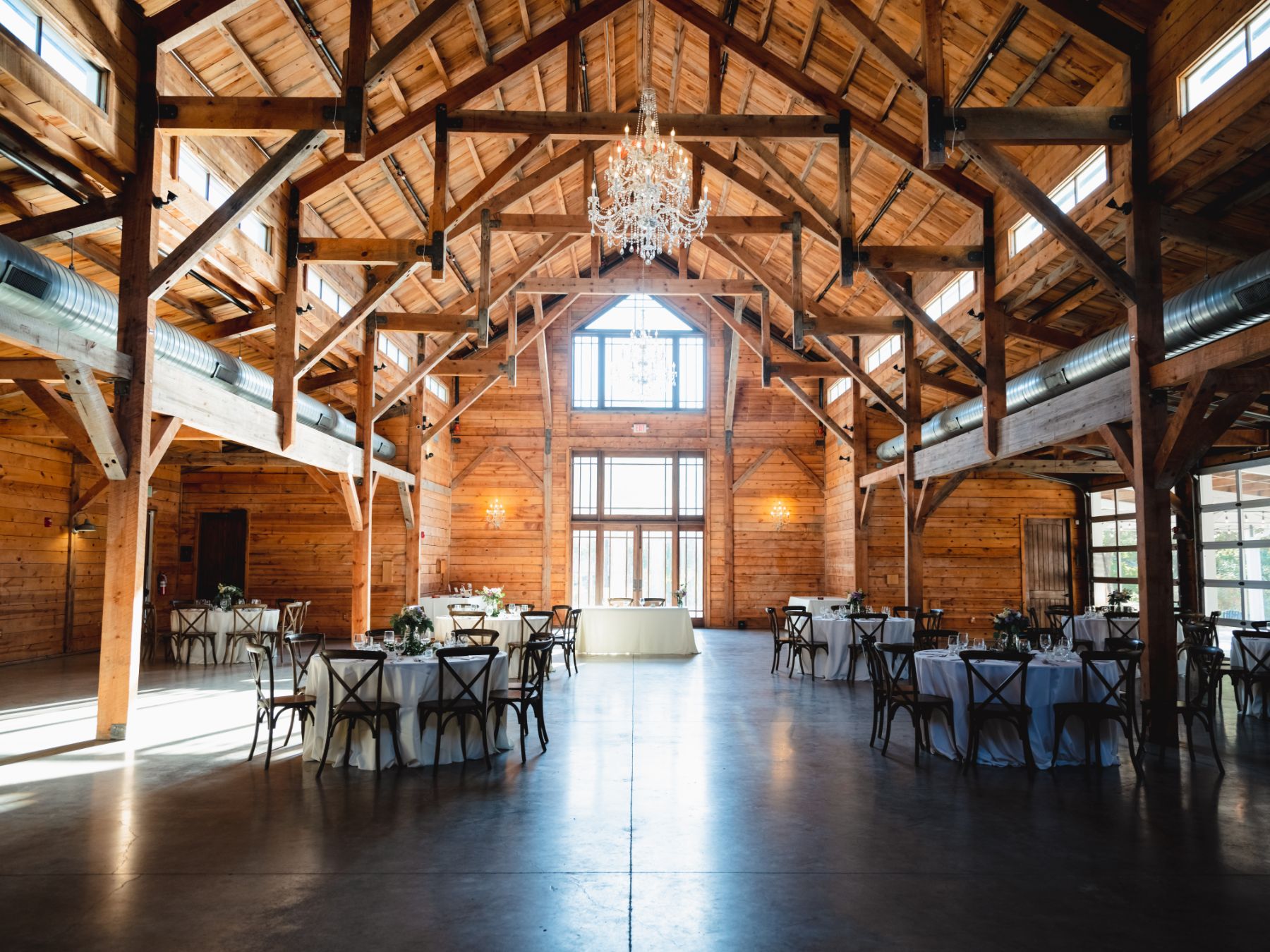 I'm inside of a wooden farm with exposed wood, walls and beans, lots of bright light, peering through large windows, and a stunning crystal chandelier at the Addison Grove wedding venue in Austin Texas.