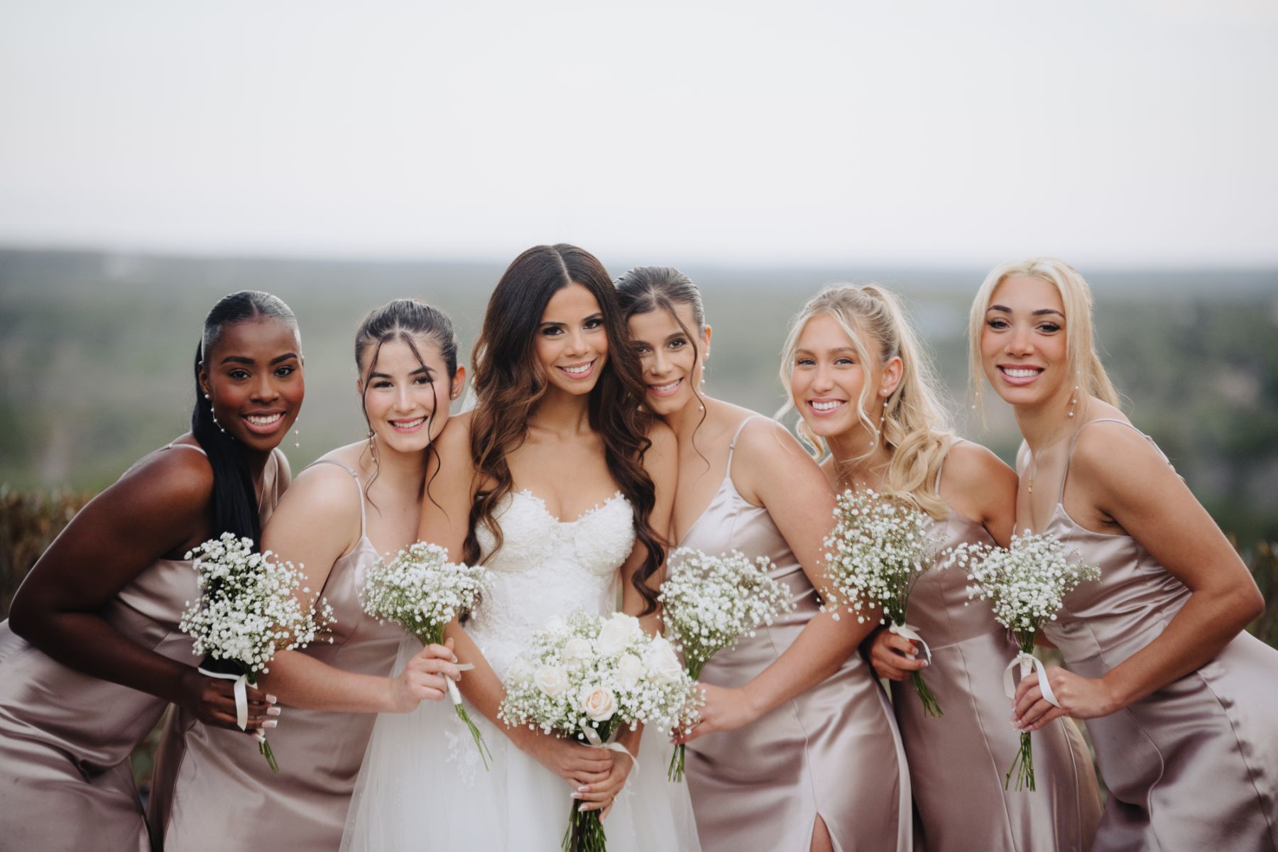 Five bridesmaids wearing blush pink silk long dresses, surround a bride with the Texas Hill country as the landscape in the background.