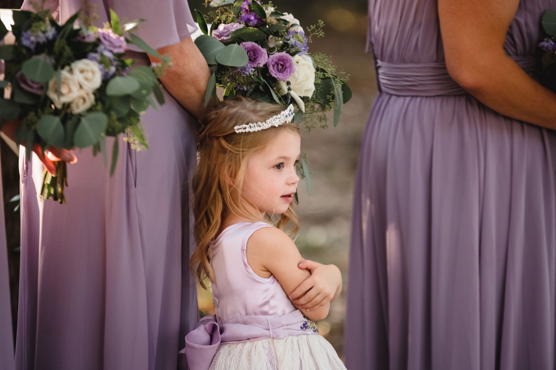 A flower girl crosses her arm, wearing a purple silk dress as she stands next to bridesmaids, wearing purple satin dresses with purple and white flower bouquets at the Addison Grove wedding venue..