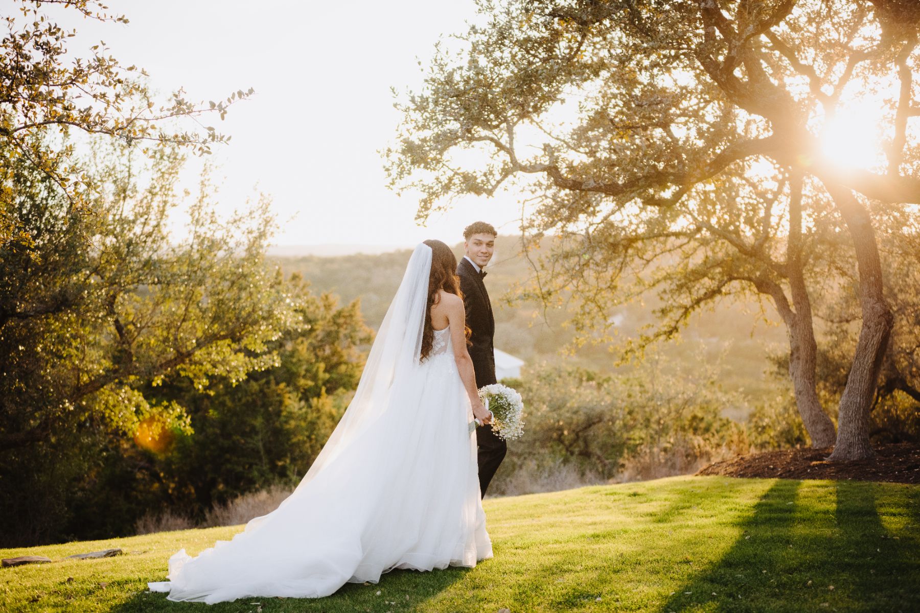 Bride and groom, walk, holding hands toward a large oak tree with the sun rays peeking through the tree during sunset at Canyonwood Ridge wedding venue.