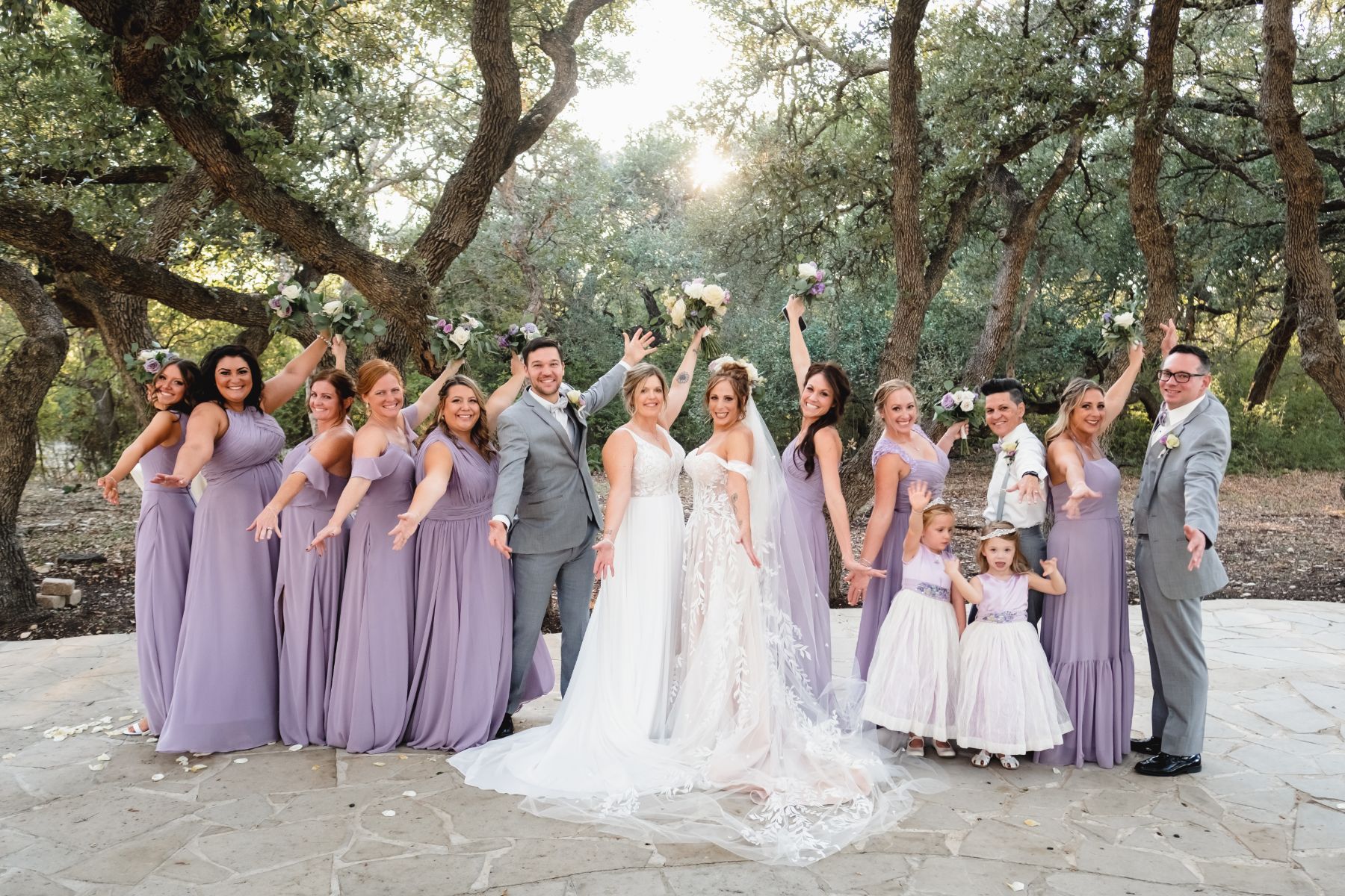 I ate bridal party, raises their hands in celebration surrounding two brides with large oak trees in the background.