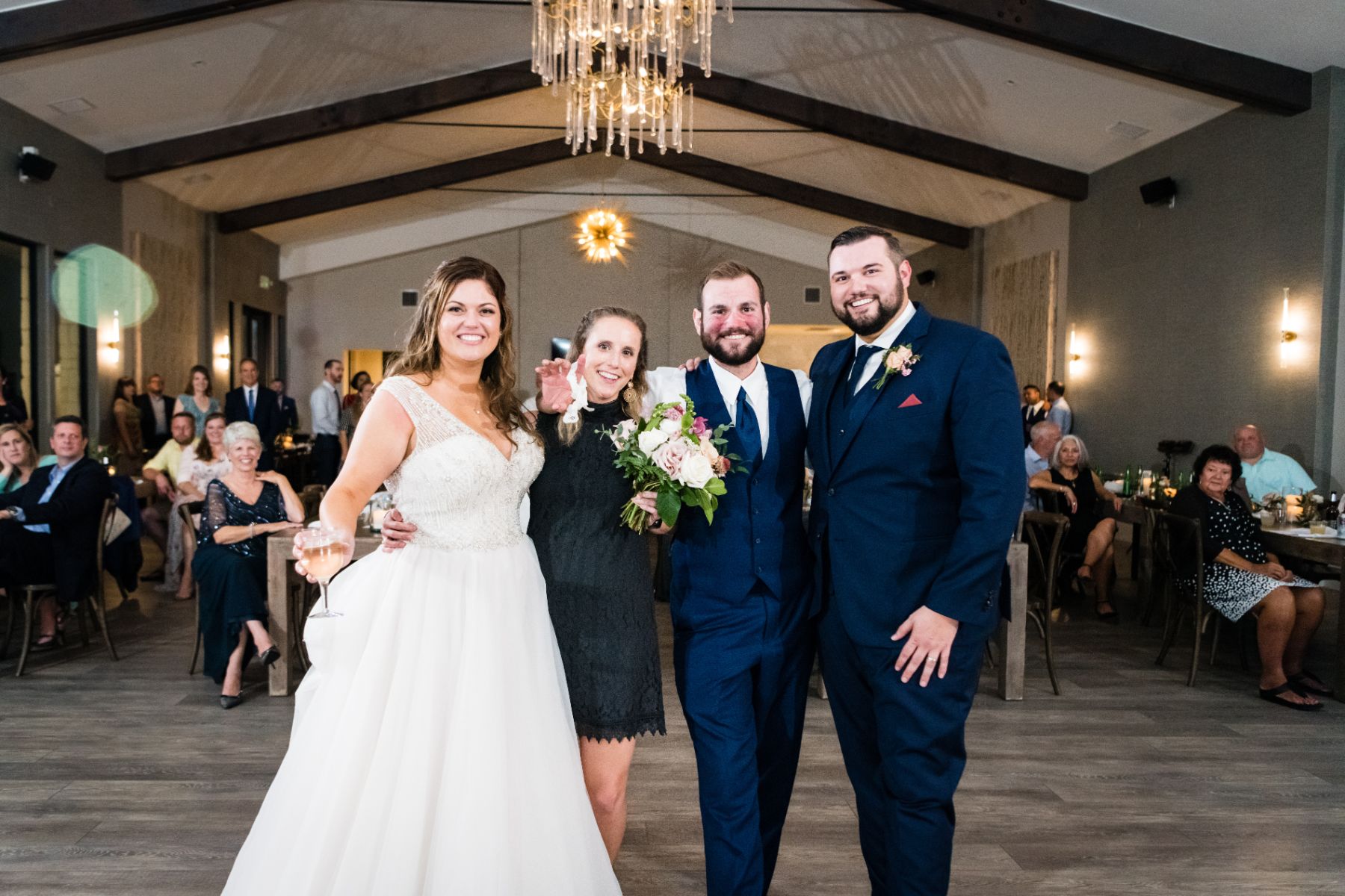 Bride and groom pose with two wedding guests during their wedding reception inside a large reception hall with exposed wood, beams, and chandeliers above at Stonehouse Villa.