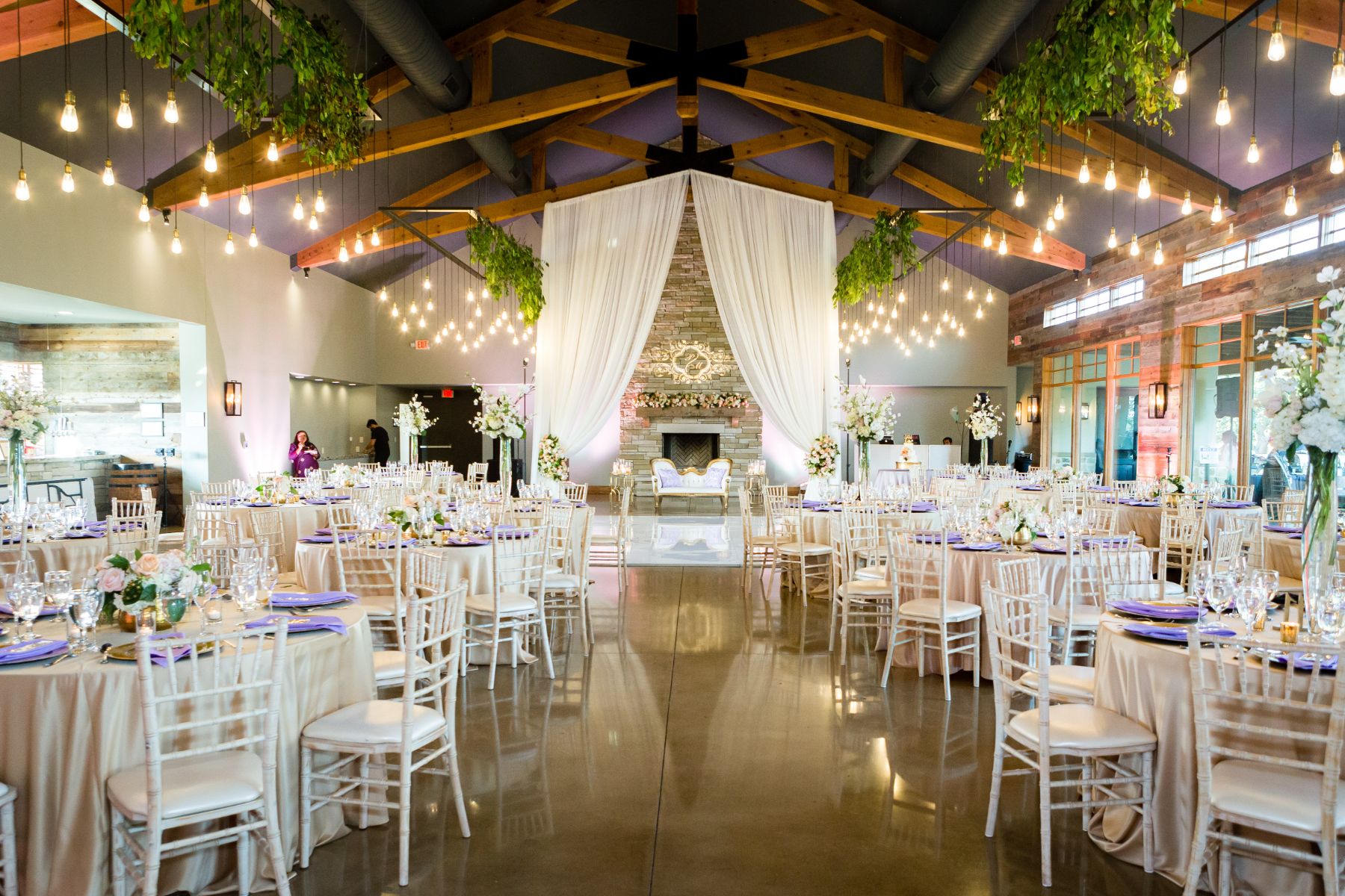 A Canyonwood Ridge wedding reception is set up in an extravagant banquet hall with wood, exposed beans and drop down pendant. Bistro lights. The tables have pink and purple color accents.