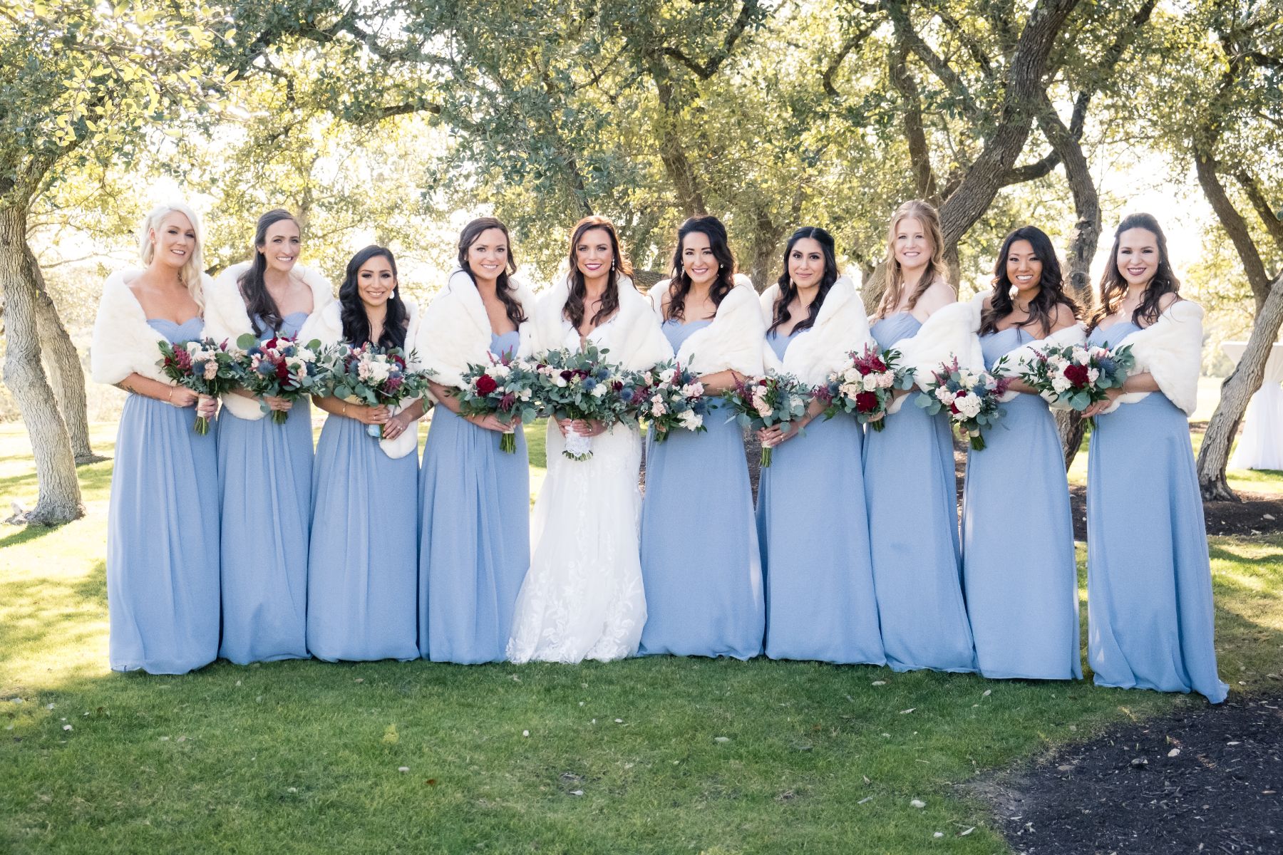 Nine. Bridesmaids wearing blue long dresses and white for shawls surround a bride. The bride and bridesmaids are standing in a line with oak trees behind them at Canyonwood Ridge Wedding venue.