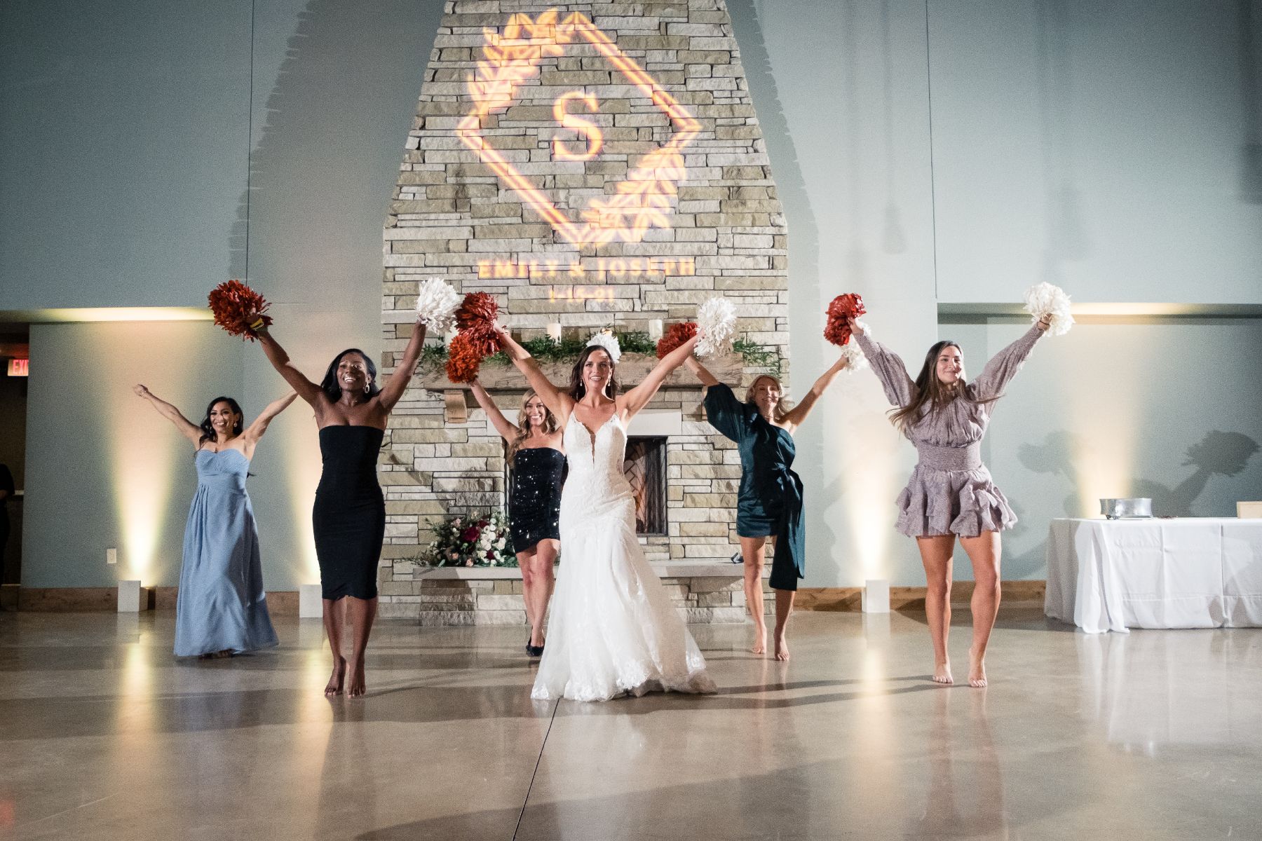 A bride stands with five women holding pom-poms in the air for a cheer. A large stone fireplace is behind them with a lit up monogram that says Emily and Joseph.
