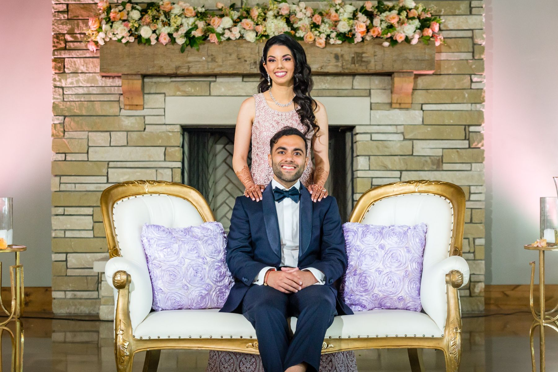 A bride stands behind a groom, sitting on a vintage, white and gold couch with a stone fireplace in the background at a Canyonwood Ridge Wedding.