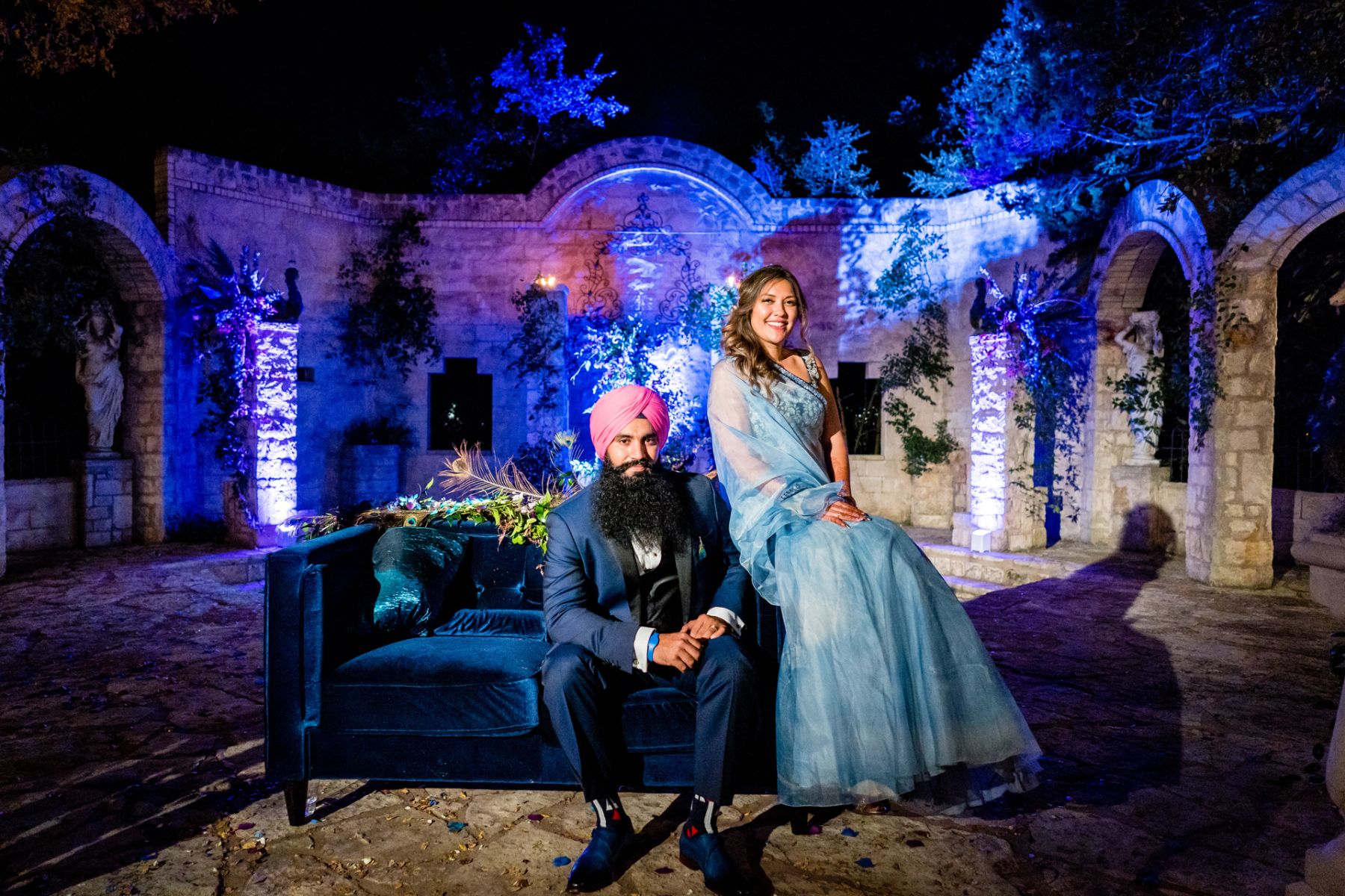 Bride and groom wearing cultural wedding garments sit on a blue velvet couch in the courtyard of The Vistas on Seward Hill with the LED lights illuminating the stone walls in the background.