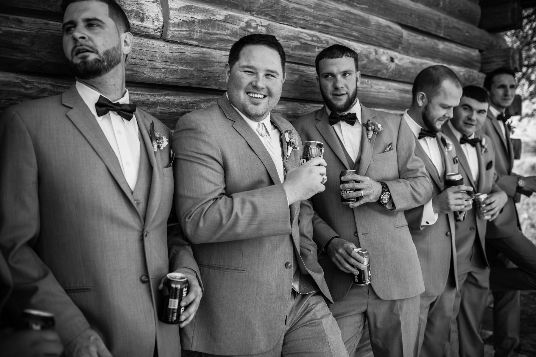 Groom and groomsmen drink beverages and smile while leaning against a rustic wood wall.